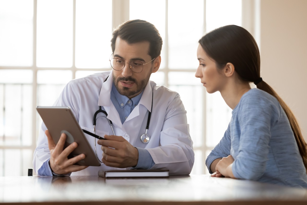 Junger dunkelhaariger Arzt zeigt einer jungen weiblichen Patientin Informationen auf seinem Tablet