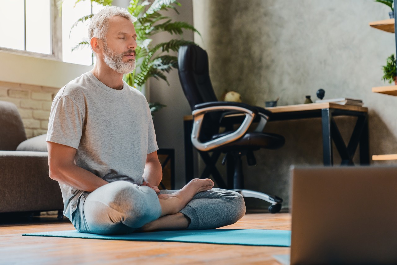 Ein Mann mittleren Alters mit grauer Kurzhaarfrisur und grauem Bart sitzt im Schneidersitz auf einer Yogamatte auf dem Boden. Seine Augen sind geschlossen und er scheint zu meditieren