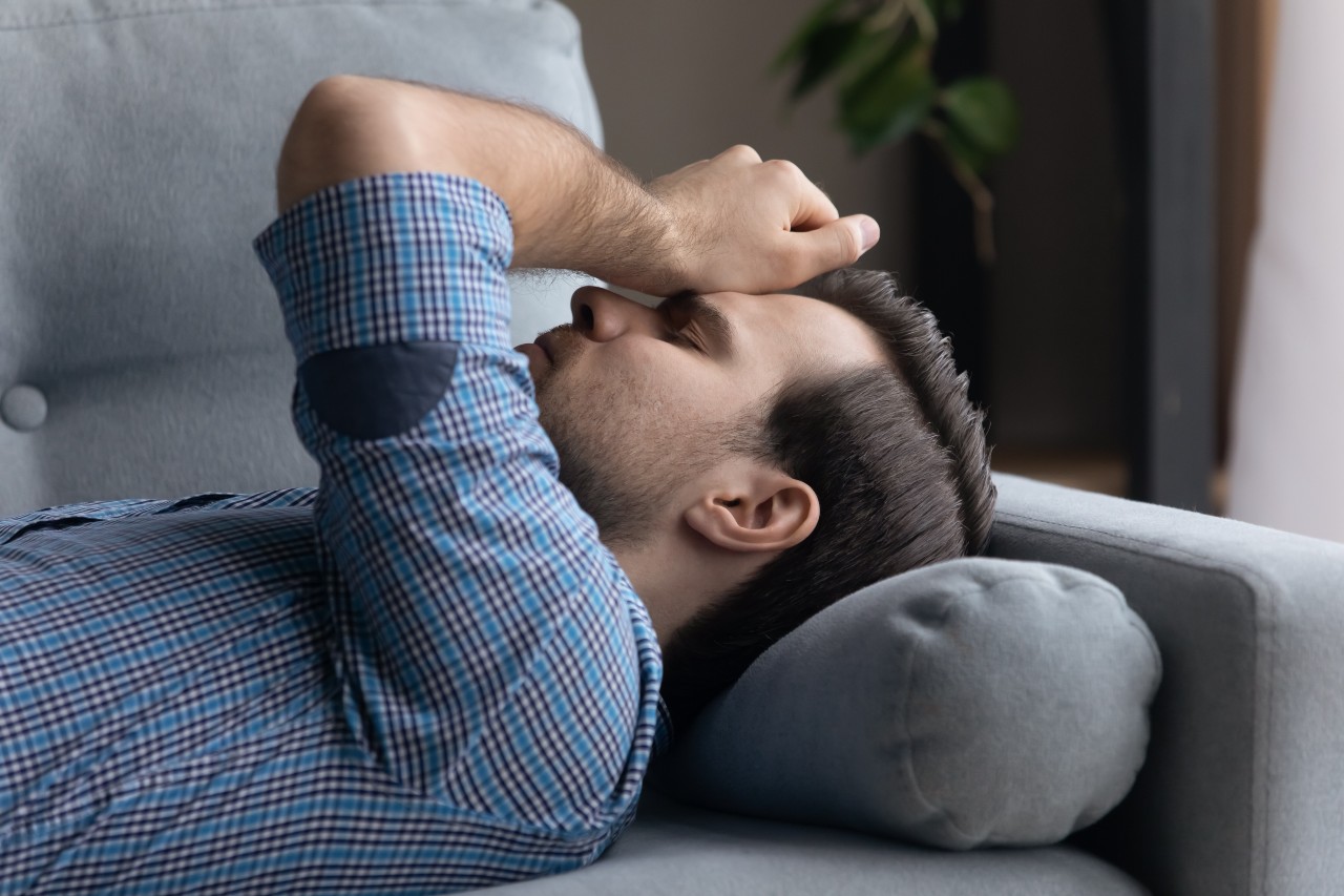 Ein junger Mann liegt rücklings auf einer Couch und fasst sich mit der linken Hand mit vor Schmerzen zusammengekniffenen Augen an die Stirn.