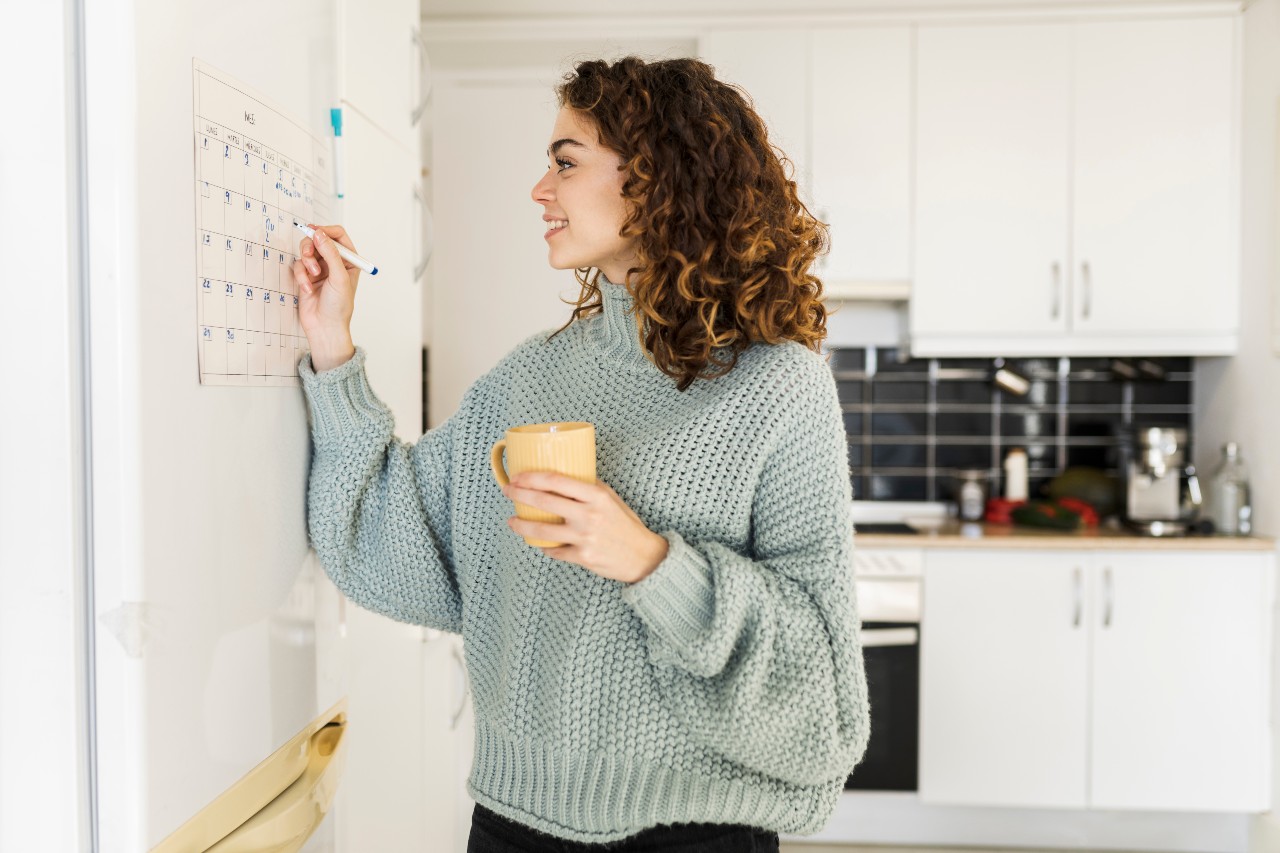 Junge Frau steht mit einem Kaffee in der linken Hand an einem Whiteboard und schreibt mit dem Stift in der rechten hand etwas darauf