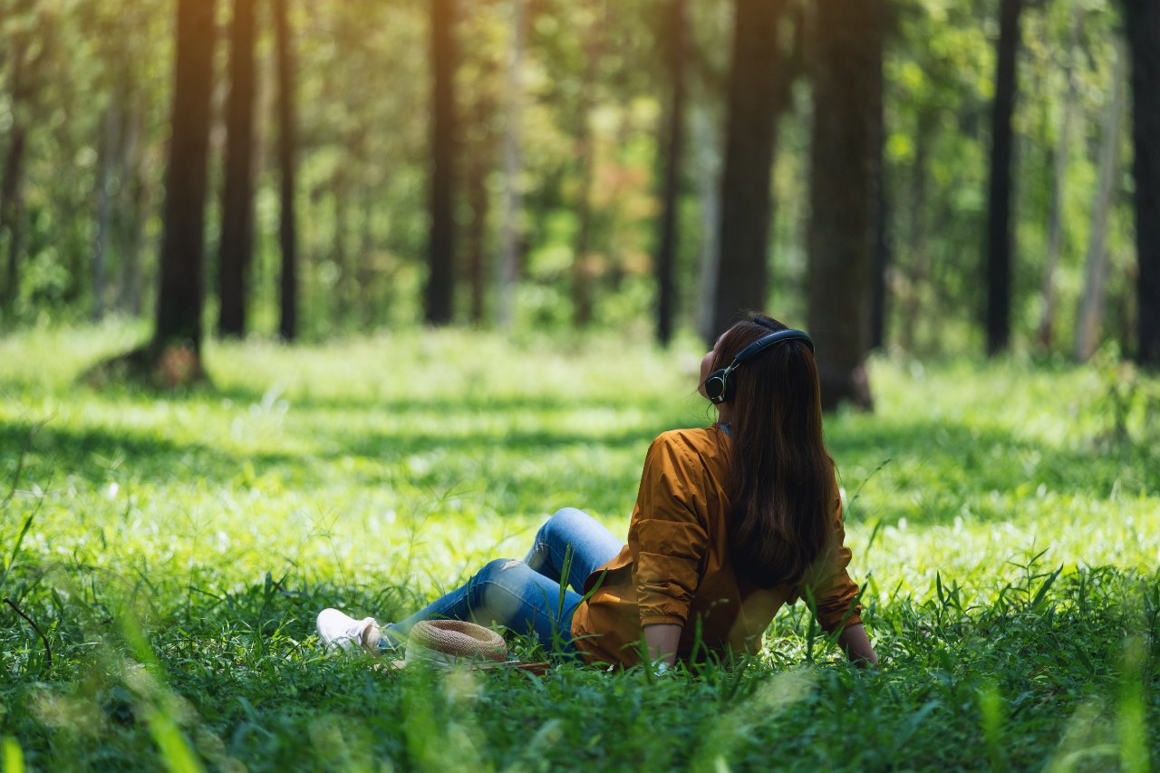 Eine junge Frau sitzt auf einer grünen Lichtung. Im Hintergrund sind Bäume zu erkennen. Sie trägt Kopfhörer und stützt sich mit den Armen auf dem Boden ab.1