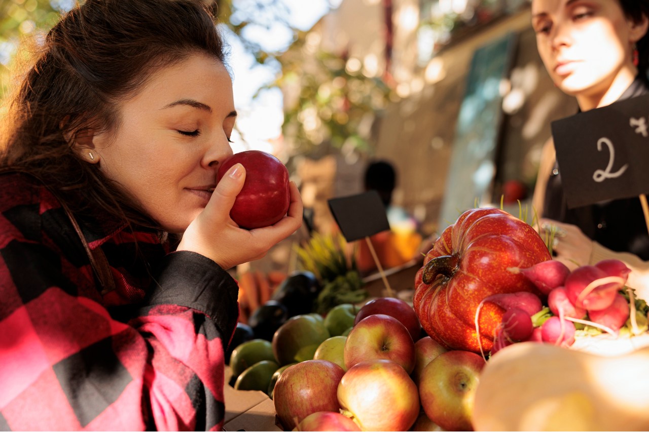 Eine Junge Frau steht an einem Obststand und riecht genussvoll an einem Apfel