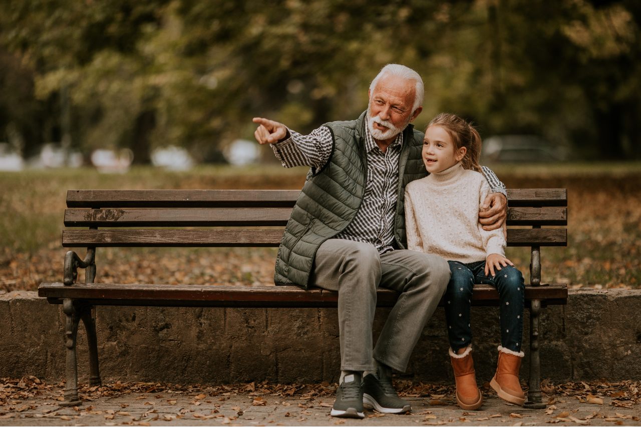 Ein Großvater sitzt mit seiner Enkelin auf einer Parkbank und zeigt mit dem Finger auf etwas außerhalb des Bildes