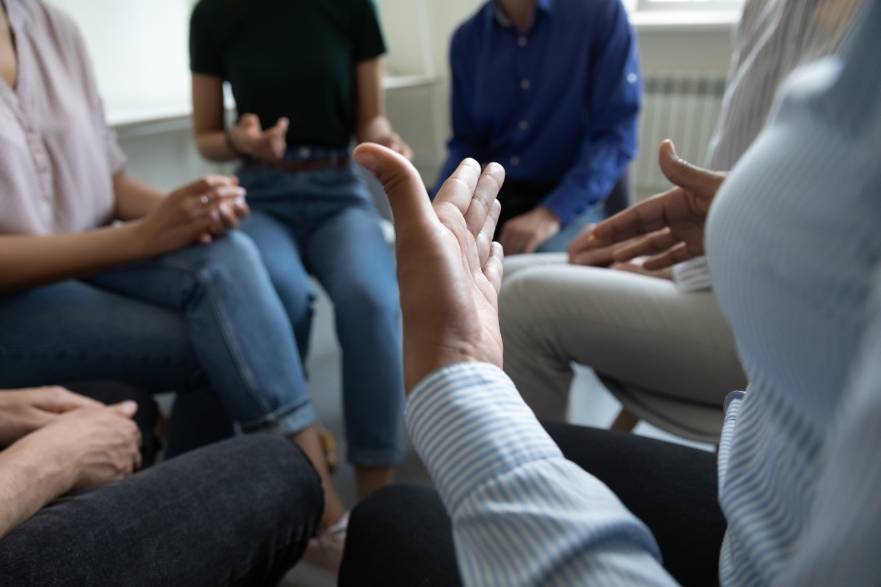 Eine Gruppe Menschen sitzt in einem Stuhlkreis. Im Vordergrund ist die Hand einer Person zu erkennen, die den anderen durch Gestik etwas mitteilen möchte.